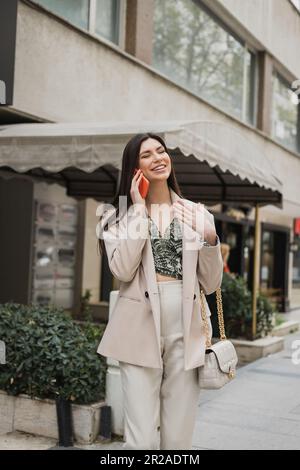 jeune femme souriante avec des cheveux longs brunette et du maquillage souriant tout en parlant sur un smartphone et debout dans une tenue tendance avec sac à main sur la sangle de chaîne n Banque D'Images