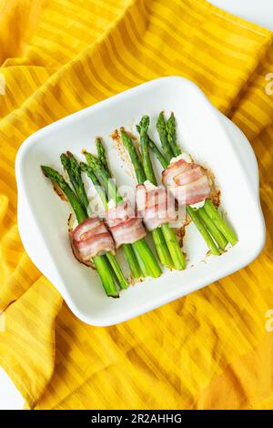 Asperges cuites au bacon et diverses épices. Un fond lumineux, une serviette orange sur laquelle repose le plat. Photo verticale Banque D'Images