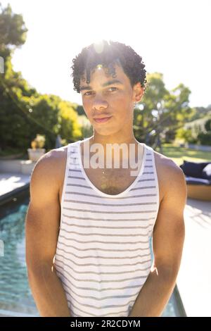 Portrait d'un homme en forme de biracial heureux portant une veste debout près de la piscine dans un jardin ensoleillé Banque D'Images