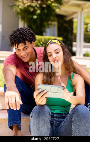 Jeune femme biraciale souriante prenant selfie avec un petit ami tout en étant assis sur des marches à l'extérieur de la maison Banque D'Images