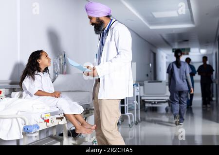 Joyeux sikh médecin mâle dans le turban et biracial fille patient parlant dans le couloir de l'hôpital, espace de copie Banque D'Images