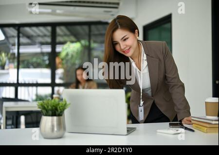Femme d'affaires ou banquier asiatique du millénaire attirante et professionnelle se penchant sur une table et utilisant son ordinateur portable à son bureau. Banque D'Images