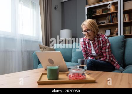 Inquiète moyenne d'âge 70s femme assis sur le canapé dans la salle de séjour lire des informations négatives inattendues sur ordinateur portable, femme adulte anxieux se reposer sur le canapé se sentent affligé wi Banque D'Images