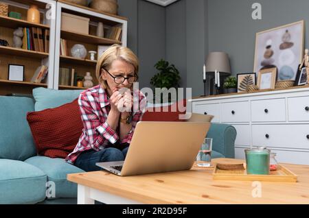 Inquiète moyenne d'âge 70s femme assis sur le canapé dans la salle de séjour lire des informations négatives inattendues sur ordinateur portable, femme adulte anxieux se reposer sur le canapé se sentent affligé wi Banque D'Images