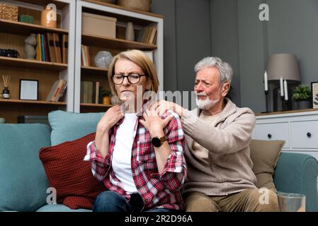 Portrait d'un couple âgé dans la salle de séjour, femme mature avec douleur dans l'épaule. Douleur au bras supérieur, douleur au cou et à l'épaule de la femme et blessure du dos elle Banque D'Images