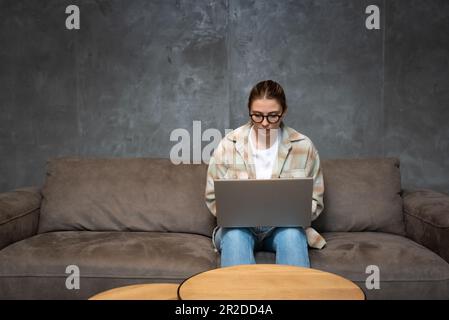 Femme indépendante ciblée utilisant un ordinateur portable à la maison, regardant l'écran, bavardant, lisant ou écrivant un e-mail, assis sur un canapé, étudiante sérieuse faisant h Banque D'Images