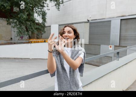 Jeune femme brune insouciante avec chandail gris chaud sur les épaules ayant appel vidéo sur smartphone tout en se tenant dans la rue urbaine avec construction industrielle Banque D'Images