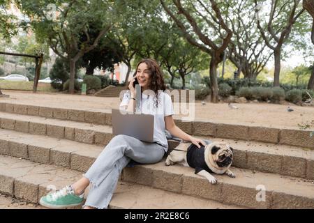 Gai brunette freelance dans des vêtements décontractés parlant sur un smartphone près d'un ordinateur portable et petting chien de pug sur les escaliers tout en passant du temps dans le parc flou dedans Banque D'Images