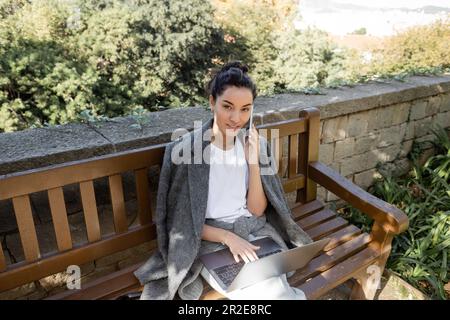 Jeune femme souriante en veste chaude parlant sur un smartphone et regardant l'appareil photo tout en travaillant sur un ordinateur portable et assis sur un banc en bois dans le parc de Barcelon Banque D'Images