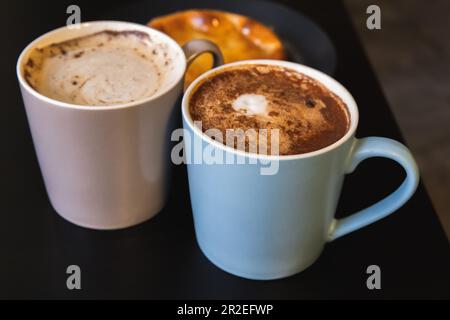 Deux Cappuccino. Les grandes tasses pleines de café avec mousse de lait et poudre de cannelle se tiennent sur une table noire, photo de gros plan avec une mise au point douce sélective Banque D'Images