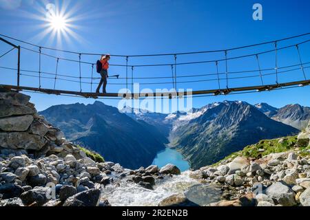 Une femme marche sur le pont suspendu au-dessus du ruisseau, Schlegeisspeicher et Großer Möseler en arrière-plan, Olpererhütte, Peter-Habeler-Runde, Zillertal Alp Banque D'Images