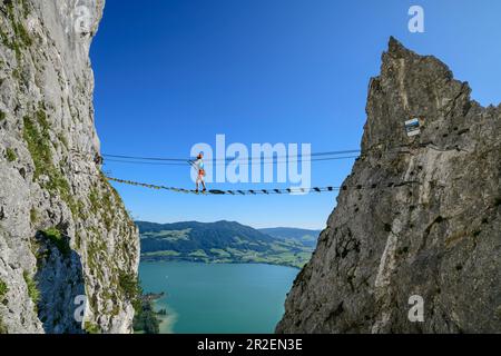 Une femme marche sur la via ferrata au-dessus du pont suspendu, via ferrata Drachenwand, Mondsee en arrière-plan, Drachenwand, Mondsee, Salzkammergut, Salzbourg, Banque D'Images