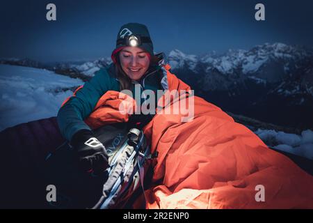 Une jeune femme se tient debout dans son sac de couchage et prend quelque chose de son sac à dos à la lumière de son phare, Karwendel, Tyrol, Autriche Banque D'Images