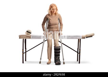 Femme devant une table médicale avec un corset de marche et un collier de cou essayant de marcher avec une béquille isolée sur fond blanc Banque D'Images