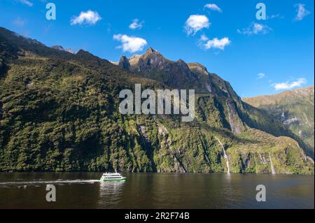 Un bateau d'excursion glisse sur une eau douce avec une toile de fond de montagnes hautes et accidentées et de cascades, Milford Sound, Parc national de Fiordland, South Island, N Banque D'Images