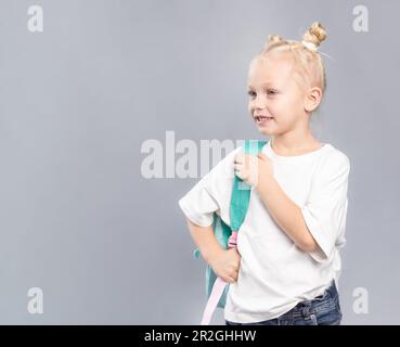 École primaire fille souriant et tenant sac à dos, enfant avec des cheveux blonds et salissants petits pains, vêtu d'un t-shirt blanc regardant sur les côtés sur le dos gris Banque D'Images