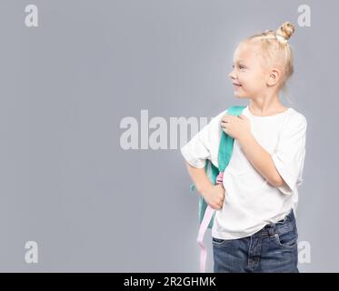 École primaire fille avec cheveux blonds et pains salissants, tenant sac à dos et vêtu de t-shirt blanc regardant latéralement sur fond gris Banque D'Images