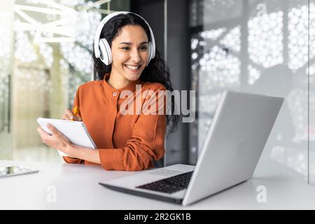 Femme d'affaires latino-américaine avec des cheveux bouclés et des écouteurs regardant le cours de formation en ligne sur le lieu de travail, femme écrivant l'information heureuse et satisfaite des résultats du développement professionnel. Banque D'Images