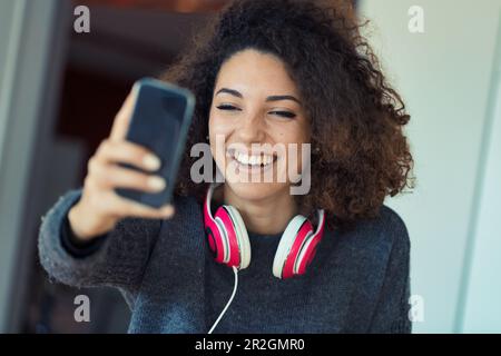 Une jeune femme aux cheveux maussés et aux pieds dans un appel vidéo. En riant et en appréciant, elle porte des écouteurs roses pour plus d'intimité. Heureux étudiant ou travailleur avec perf Banque D'Images