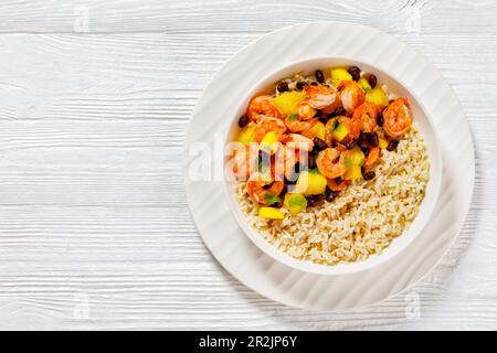 Crevettes grillées des Caraïbes aux haricots noirs, salsa à l'ananas à la mangue servie avec du riz brun dans un bol blanc sur une table en bois blanc, vue horizontale depuis le dessus Banque D'Images