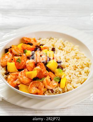 Gros plan de crevettes grillées des Caraïbes aux haricots noirs, salsa à la mangue et à l'ananas servi avec du riz brun dans un bol blanc, vue verticale Banque D'Images