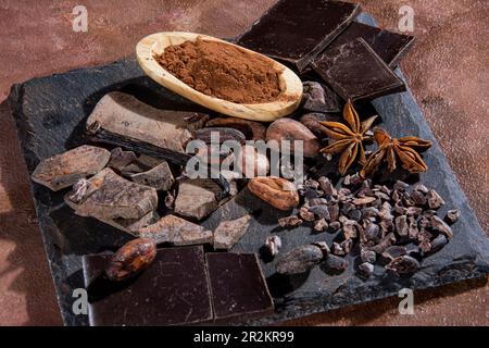 Comprimé de chocolat noir haché sur une dalle d'ardoise, accompagné de poudre de cacao, de chocolat pur, de fèves de cacao grillées et d'autres ingrédients, photographié i Banque D'Images