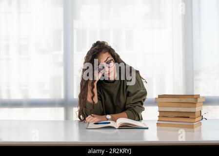 Surtravaillé. Une jeune femme fatiguée s'assoit au bureau de la maison bondé de livres cacher le visage dans les palmiers sentir mal de tête. Une femme exténuée se sent mal lo Banque D'Images