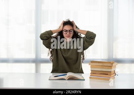 Surtravaillé. Une jeune femme fatiguée s'assoit au bureau de la maison bondé de livres cacher le visage dans les palmiers sentir mal de tête. Une femme exténuée se sent mal lo Banque D'Images