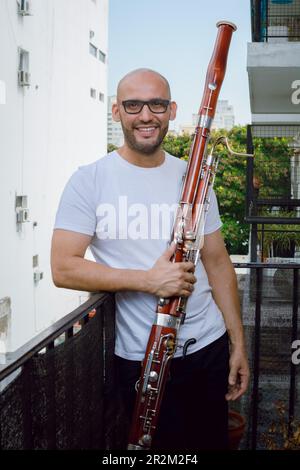 portrait vertical du jeune latino avec des lunettes et une barbe, souriant à la maison tenant son basson et souriant regardant l'appareil photo Banque D'Images