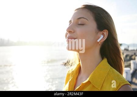 Jeune femme respirant de l'air frais avec les yeux fermés écoutant de la musique relaxante avec des écouteurs sans fil sur la baie de la ville au coucher du soleil Banque D'Images