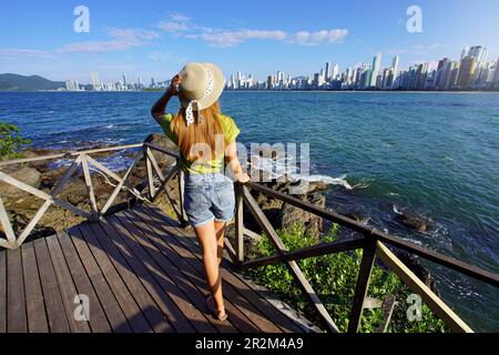 Pleine longueur de jeune femme avec chapeau profitant de l'horizon de la métropole sur la baie au coucher du soleil, Balneario Camboriu, Brésil Banque D'Images