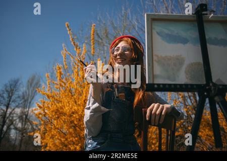 Gros plan d'une fille de gingembre souriant et peignant sur sa toile tout en étant assis au parc devant les fleurs jaunes Banque D'Images