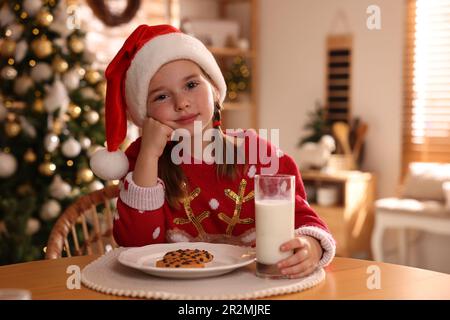 Jolie petite fille en chapeau de père Noël avec un délicieux cookie de Noël et du lait à la maison Banque D'Images