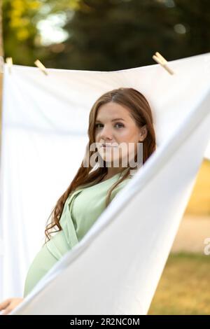 Jolie femme enceinte blanche accrochant la lessive sur la ligne de vêtements à l'extérieur, linge de lit de séchage, literie. Détergents à lessive sûrs, gel de lavage organique Banque D'Images