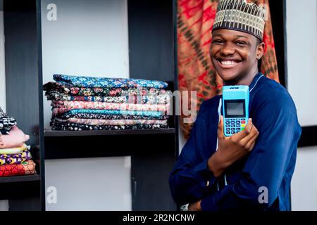 Photo d'un homme africain noir debout à côté d'une armoire en tissu tout en tenant et en payant avec une machine de pos dans un magasin de textile Banque D'Images