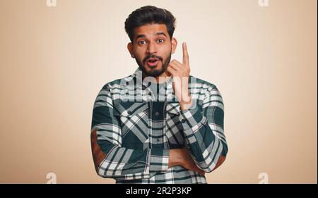 Eureka. Jeune Indien inspiré pointant le doigt vers le haut avec la bouche ouverte, ayant une bonne idée, plan, démarrage, montrant l'inspiration mouvement de motivation, solution de problème. Homme hindou barbu sur fond beige Banque D'Images