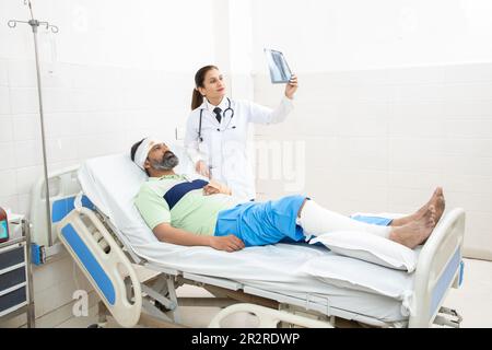 Patient indien allongé sur le lit avec une jambe fracturée et médecin orthopédique regardant le film radiographique du pied donnant des conseils de rétablissement. Banque D'Images