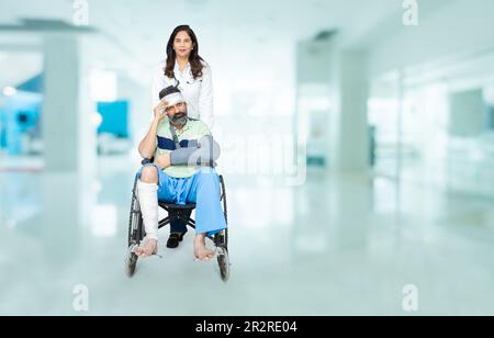 Jeune indien avec une jambe fracturée après un accident assis sur un fauteuil roulant poussée par le personnel médical à l'hôpital, concept de soins de santé.Copy space. Banque D'Images