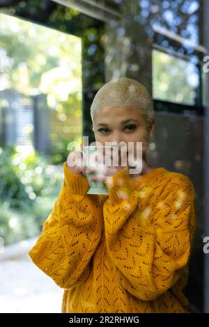 Portrait non modifié de la femme biraciale à la mode heureuse tenant une tasse de café, vue à travers la fenêtre. Détente, temps libre et style de vie domestique. Banque D'Images