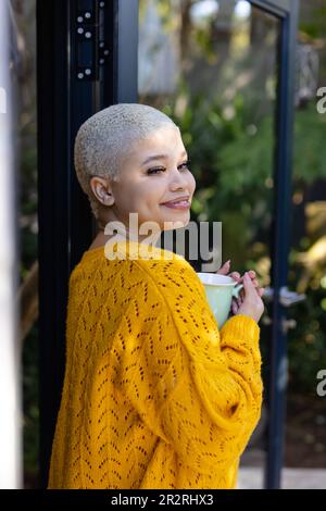 Portrait d'une femme biraciale à la mode et à la mode, tenant une tasse de café, debout près d'une fenêtre. Détente, temps libre et style de vie domestique. Banque D'Images