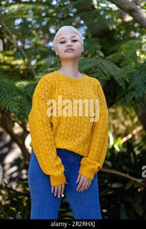 Portrait non modifié de la femme biraciale à la mode heureuse en chandail jaune debout près des arbres dans le jardin. Détente, temps libre, nature et style de vie. Banque D'Images