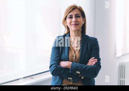 Femme d'affaires caucasienne âgée de 50s ans, dans une tenue de bureau décontractée, debout près de la fenêtre dans la salle blanche de bureau, avec les bras croisés, souriant à la caméra. Banque D'Images
