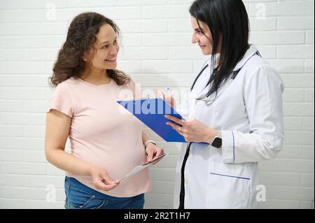 Femme médecin gynécologue avec presse-papiers consulter une femme enceinte gravid, prescrire des médicaments. Grossesse. 9 mois Banque D'Images
