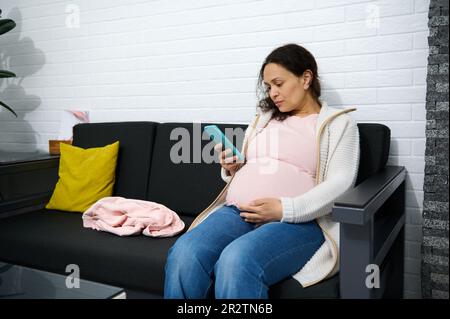 Femme enceinte vérifiant son téléphone portable tout en étant assise sur le canapé dans la salle d'attente d'une clinique médicale gynécologique Banque D'Images