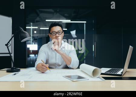 Portrait d'un jeune homme asiatique designer, architecte, ingénieur regardant avec confiance et sérieux la caméra, travaillant au bureau à une table avec des papiers et des projets. Banque D'Images