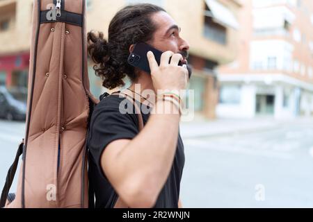 Jeune homme hispanique musicien parlant sur smartphone tenant une caisse de guitare dans la rue Banque D'Images
