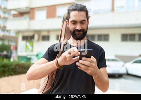Jeune homme hispanique musicien utilisant un smartphone tenant une caisse de guitare au parc Banque D'Images