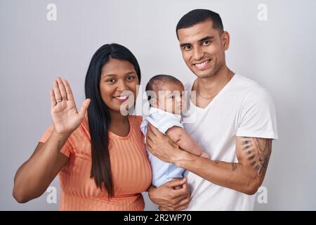 Jeune couple hispanique avec bébé debout ensemble sur un fond isolé renonçant à dire bonjour heureux et souriant, geste de bienvenue amical Banque D'Images