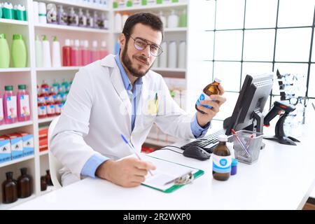 Jeune homme hispanique pharmacien tenant une bouteille de médicament écrit sur le document à la pharmacie Banque D'Images