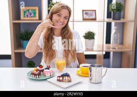 Jeune femme caucasienne mangeant des pâtisseries t pour le petit déjeuner souriant faisant un geste de téléphone avec la main et les doigts comme parler au téléphone. communication c Banque D'Images